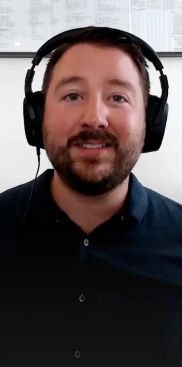 Man with a beard wearing black headphones and a dark shirt smiling indoors.